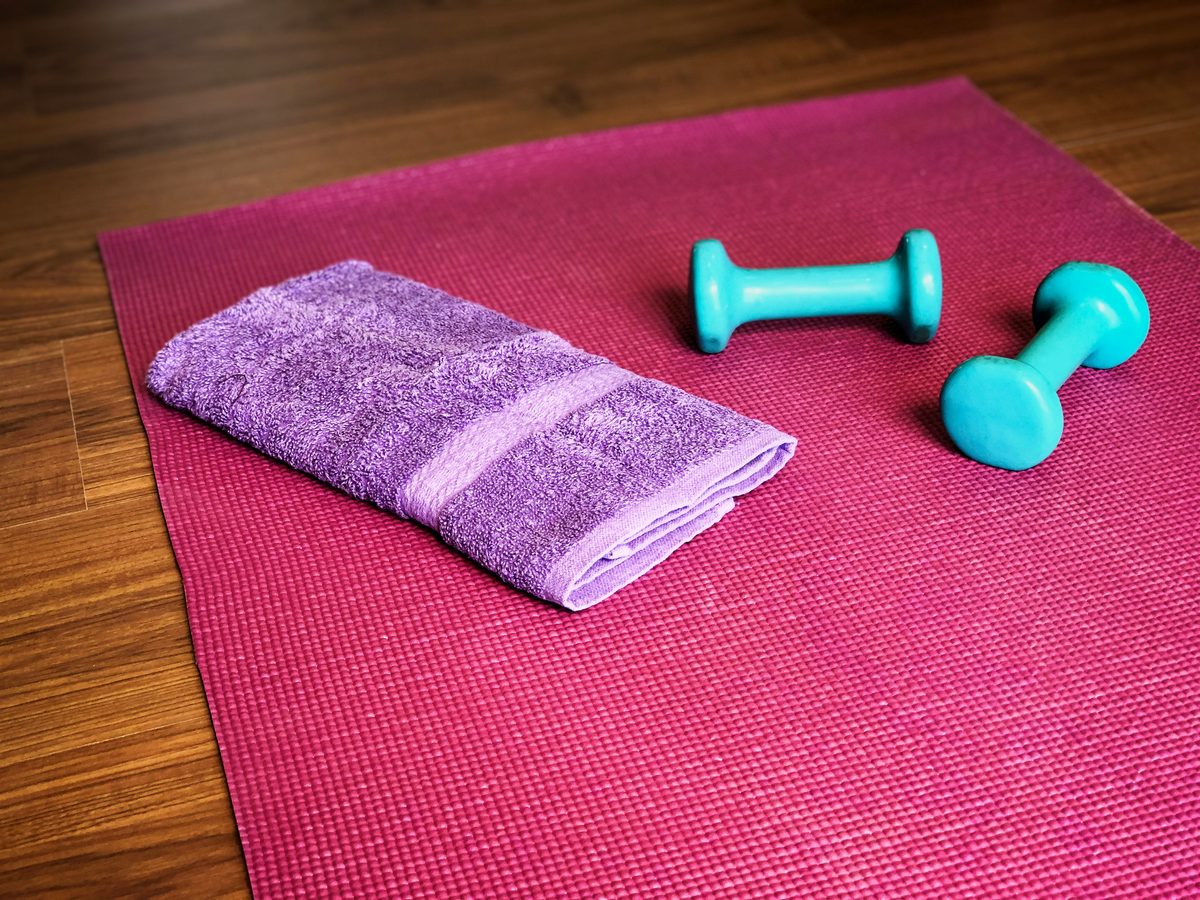An exercise mat with a folded towel and two light dumbbells on a wood floor